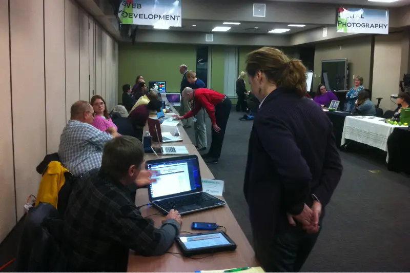 Many people gathered in a room observing various websites displayed on laptops, with signs in the background reading 'Web Development' and 'Digital Photography'